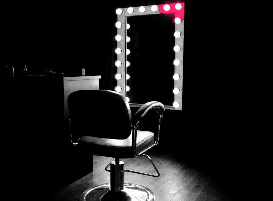An empty salon chair facing a bulb-lit mirror